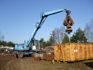 Beitragsbild: Hier wird ein Bagger auf einem Schrottplatz gezeigt der der mittels eines Schrottgreifers und eines Magneten Schrott in einem Container aussortiert.